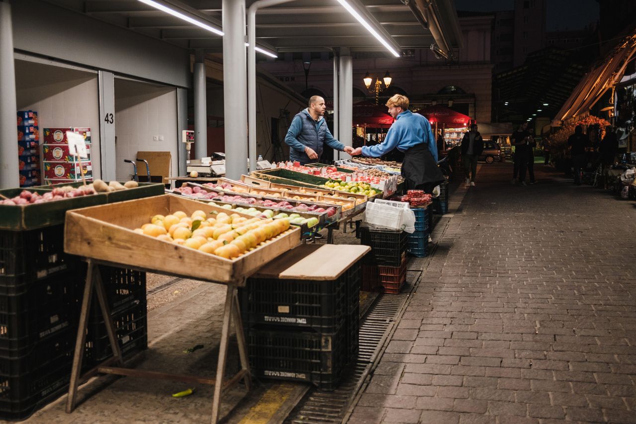 services-05 Two people exchanging goods at a well-lit night market in the city, surrounded by fresh produce.