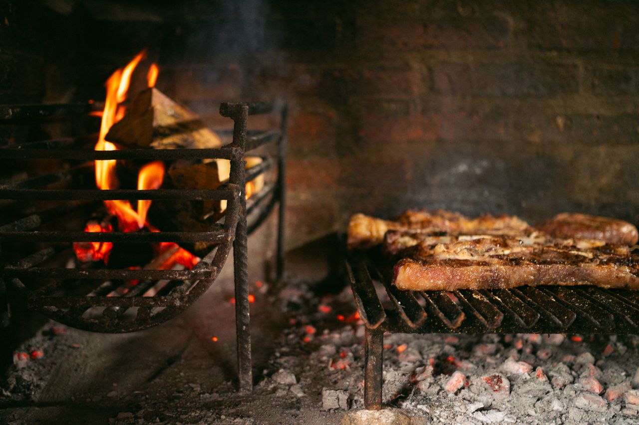 why-choose-us Juicy steaks cooking over an open fire barbecue in a cozy rustic setting.